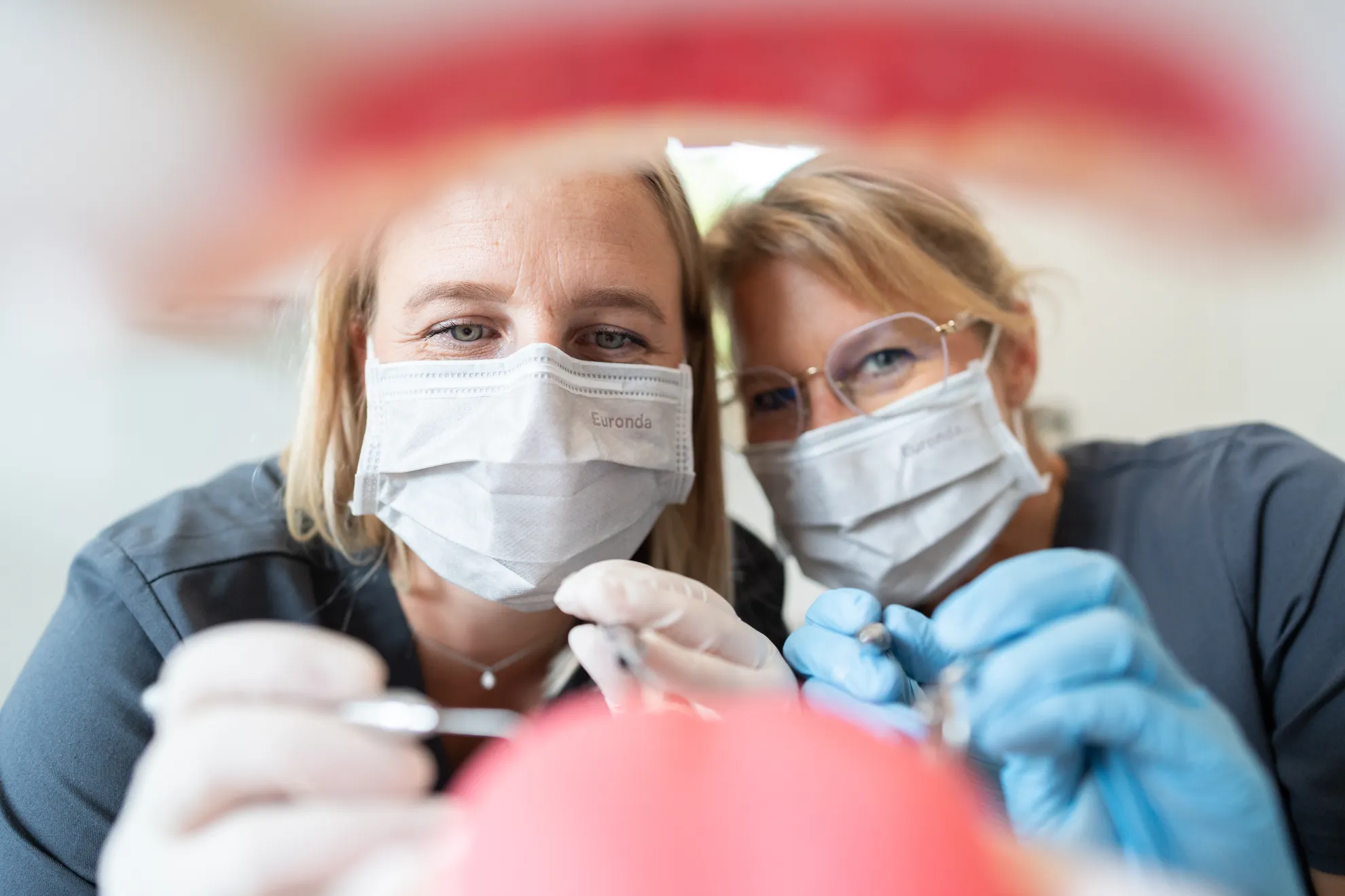 Zwei Zahnärztinnen mit Mundschutz und Instrumenten beim Blick in den Patientenmund (in dem Fall nur ein Zahnmodell aus Kunststoff)