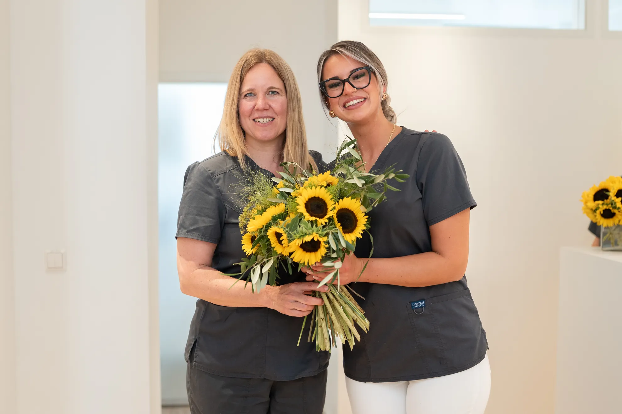 Zwei Kolleginnen mit Sonnenblumen lächelnd vor weißer Wand